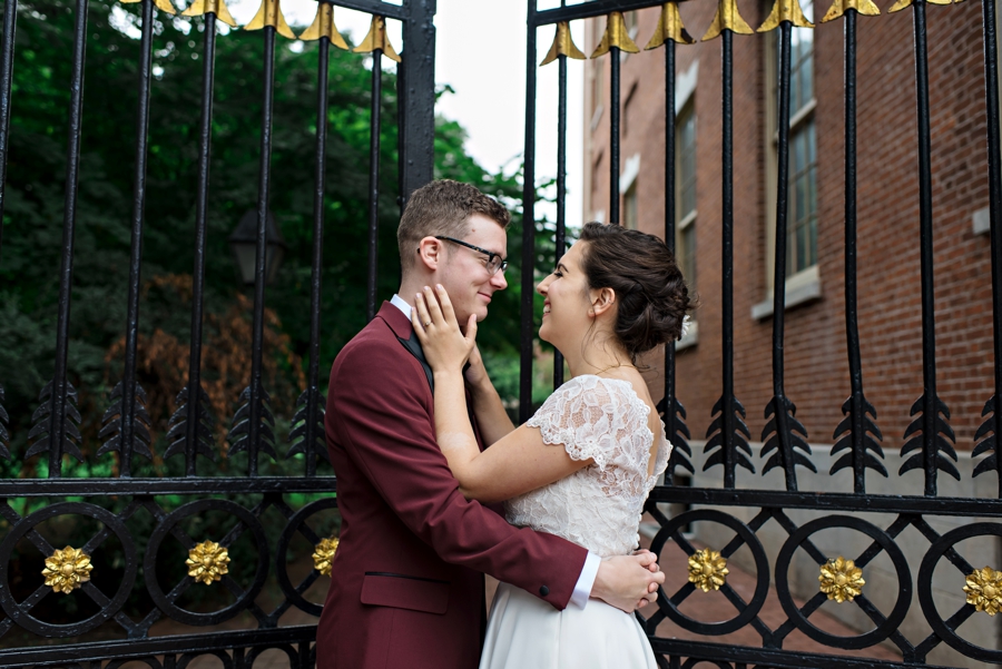 A bride holds her grooms face and smiles while standing in front of iron gates.